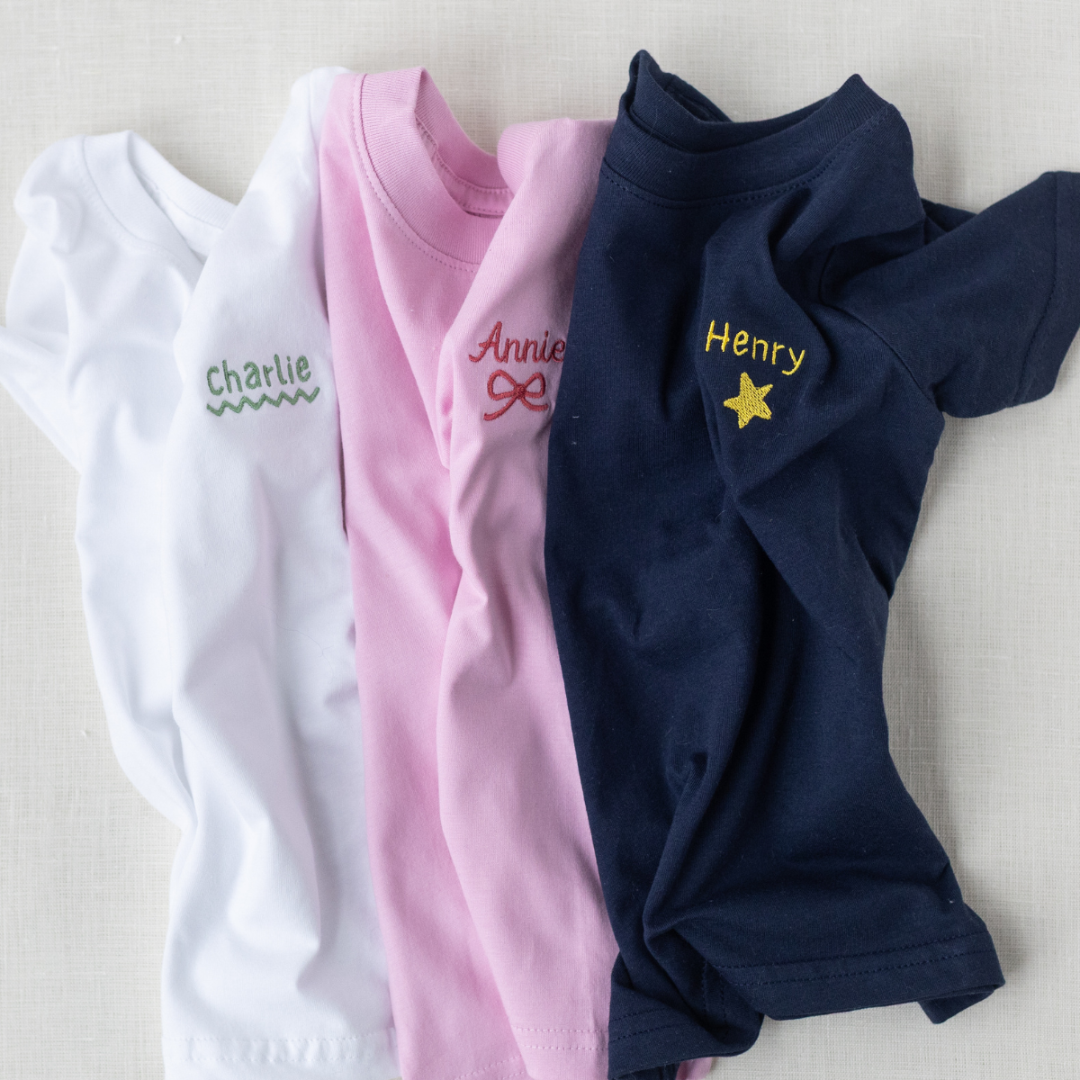 Three folded shirts in white, pink, and navy with embroidered names on a light background