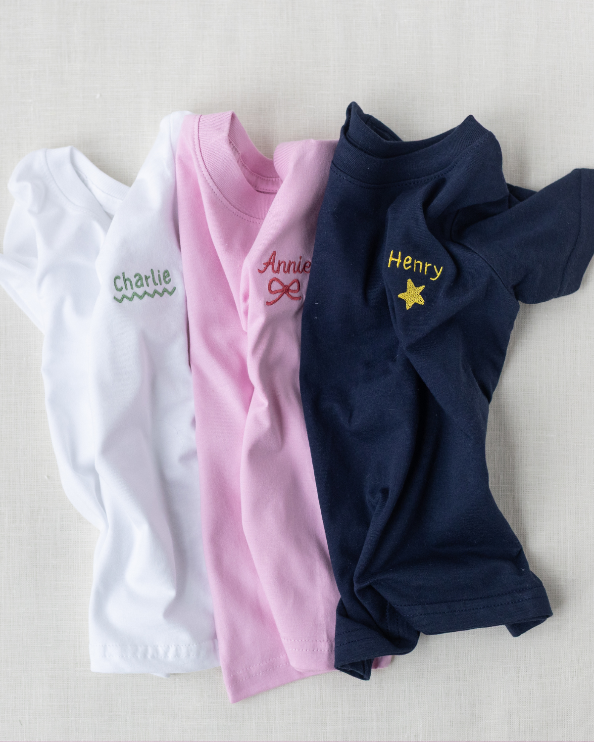 Three folded shirts in white, pink, and navy with embroidered names on a light background