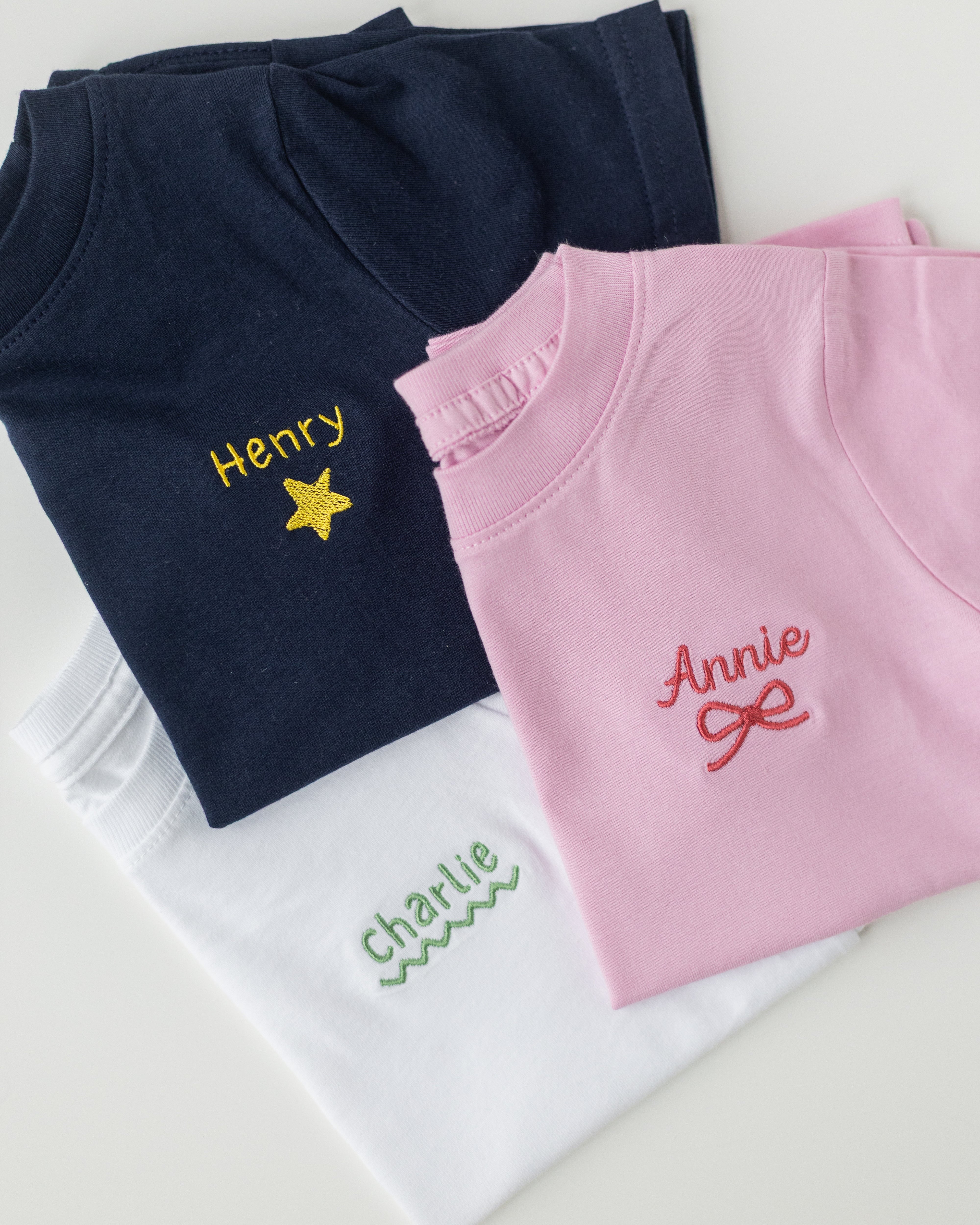 Navy, pink, and white shirts with embroidered names on a light gray background